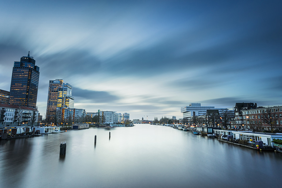 A grey sky reflects in an urban river with buildings either side bluehealth