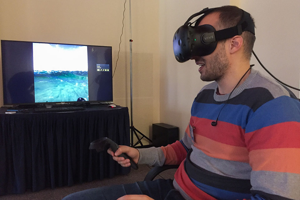 A man sits in a chair with a VR headset on - a screen behind shows a virtual beach